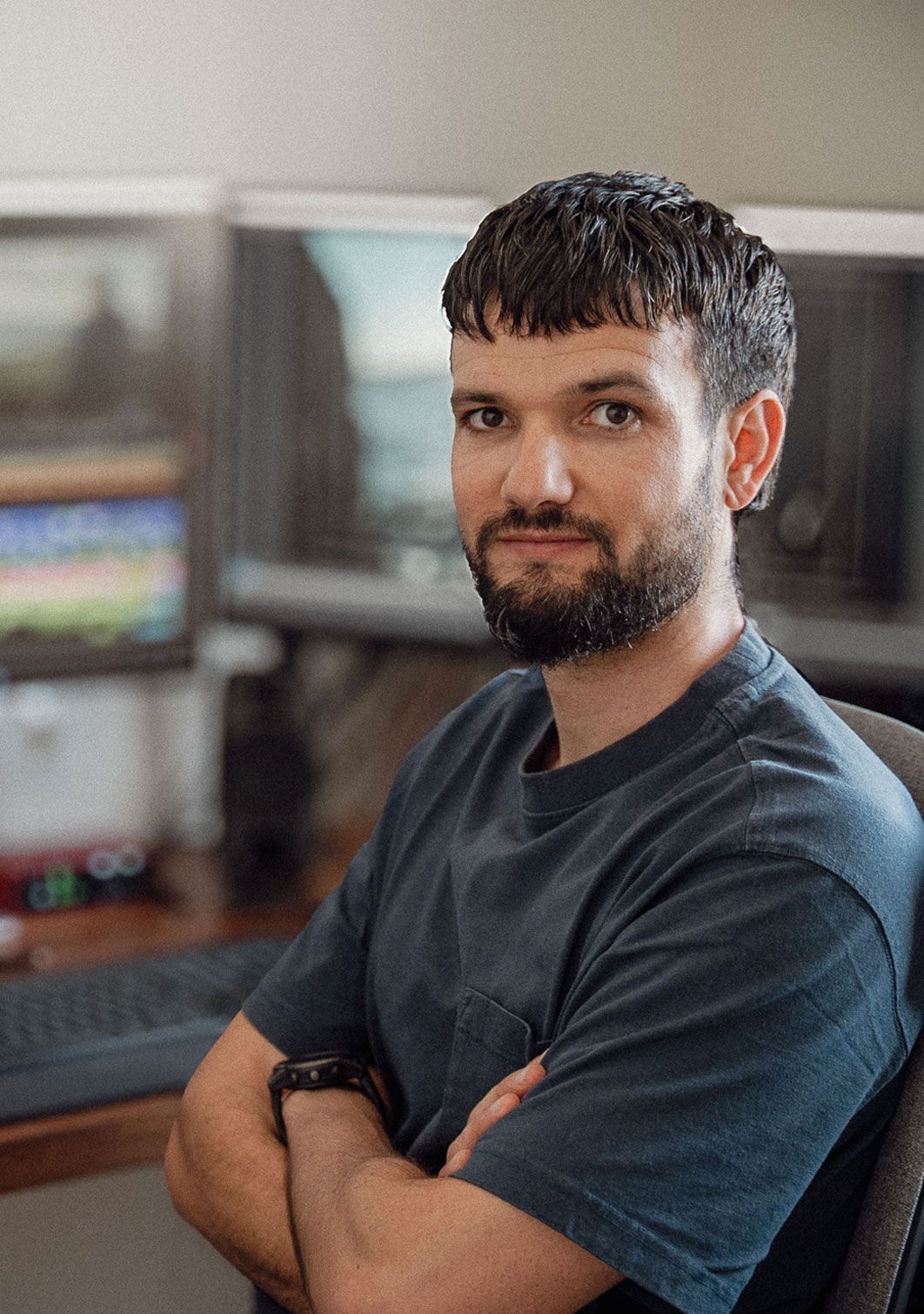 Un hombre de cabello oscuro y barba está sentado en un escritorio con los brazos cruzados, mirando a la cámara. Al fondo se ven monitores de computadora y un teclado.