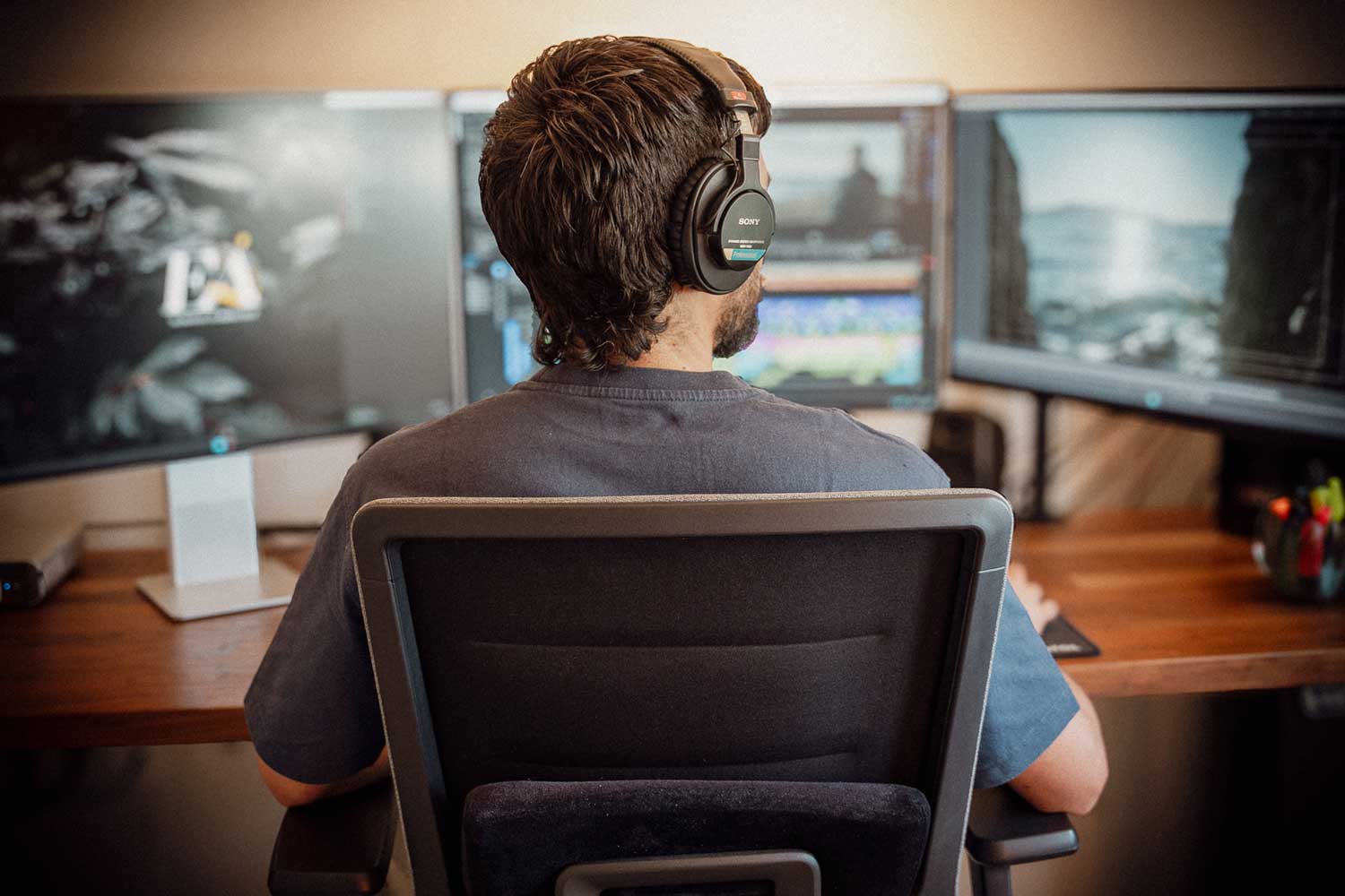 Una persona con auriculares se sienta en un escritorio con tres monitores de computadora que muestran varias imágenes y software de edición.