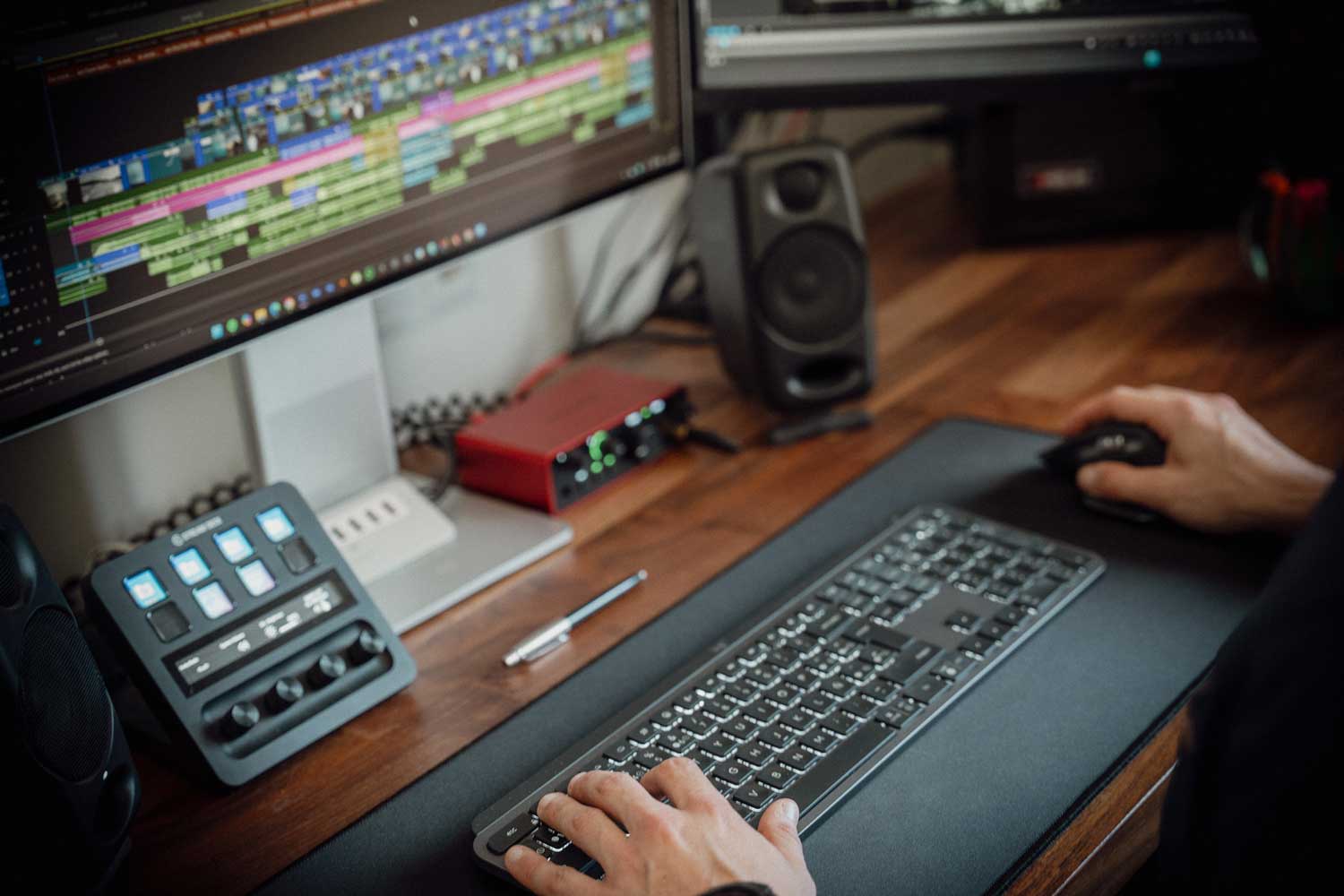 Una persona utiliza un ratón de computadora frente a un teclado y un monitor que muestra una línea de tiempo de edición de vídeo, con equipo de audio y altavoces en un escritorio de madera.